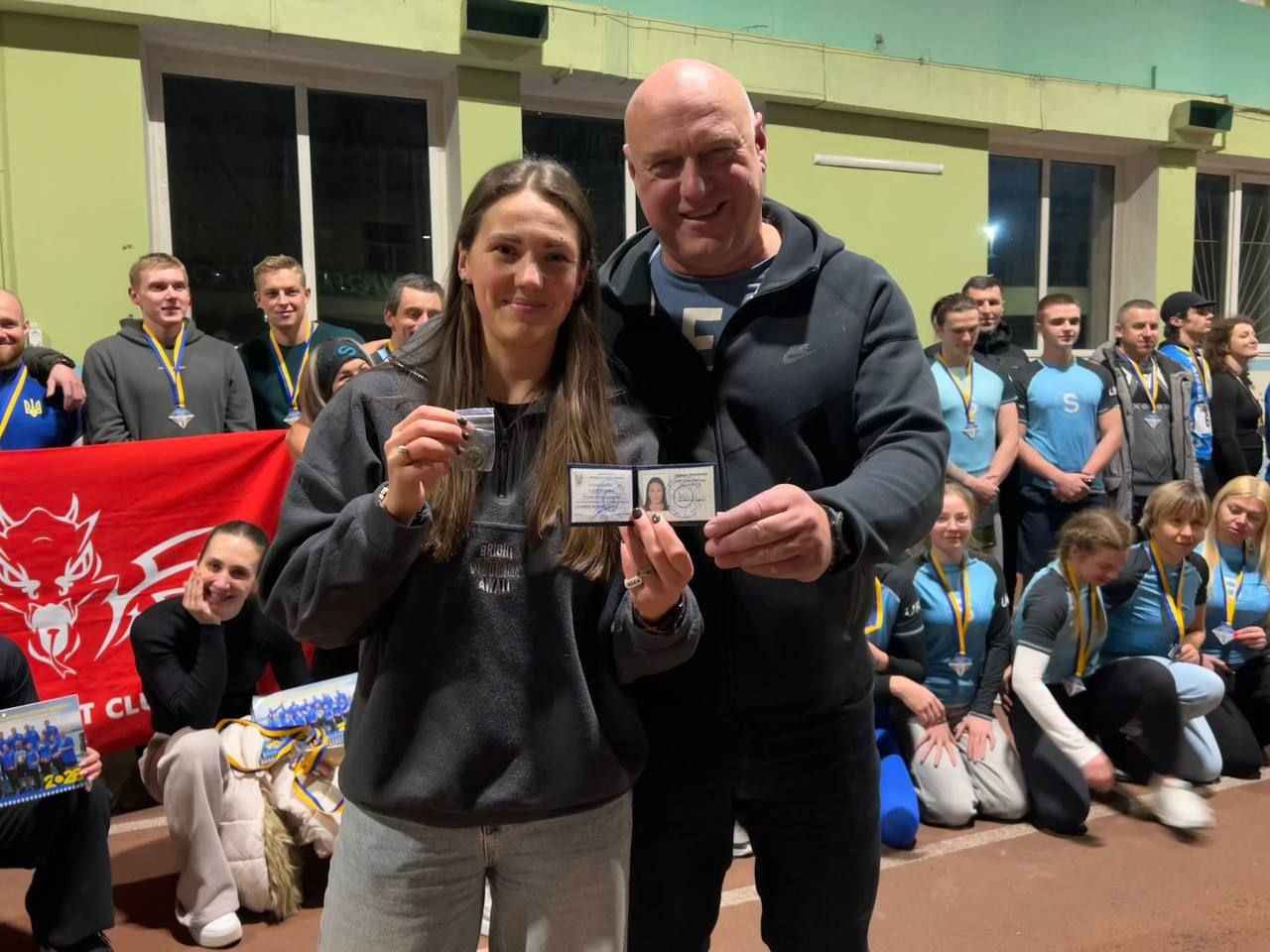 Групове фото в спортивному залі, де чоловік і жінка на передньому плані тримають спортивне посвідчення та медаль. На задньому плані стоїть група молодих людей у спортивній формі з медалями на шиях, а ліворуч видно частину червоного прапора з емблемою.