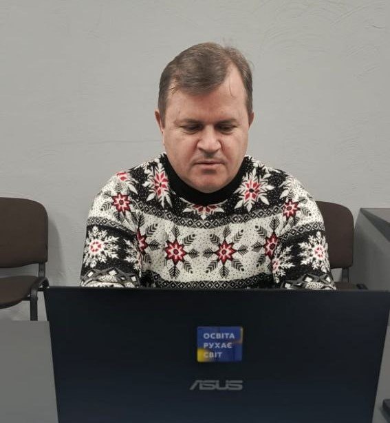 Чоловік у світлому светрі з орнаментом у вигляді сніжинок сидить за столом і зосереджено працює за ноутбуком. На кришці ноутбука помітно невелику наклейку з написом: ОСВІТА РУХАЄ СВІТ на фоні кольорів державного прапора України.