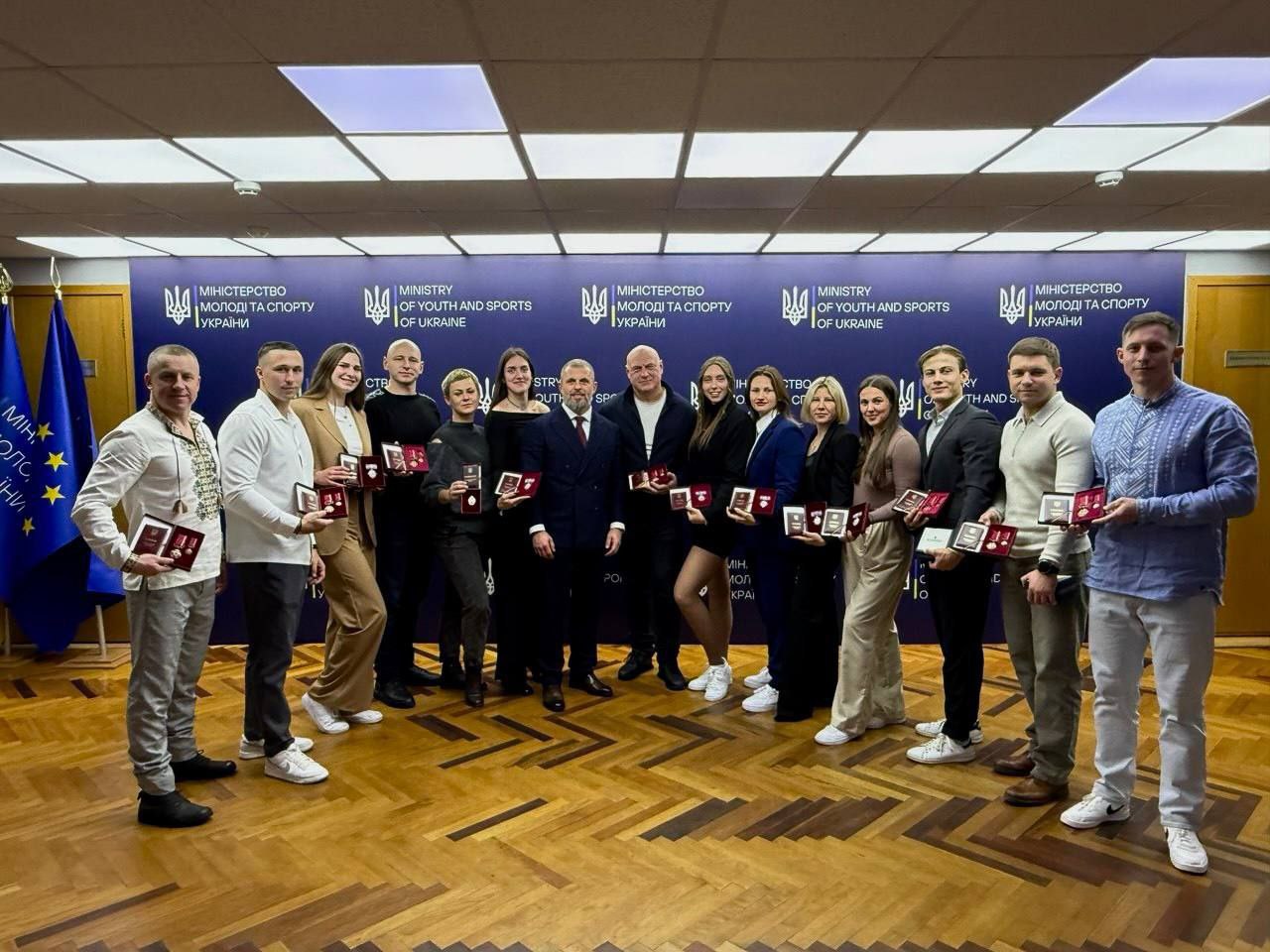 Групове фото офіційної церемонії нагородження у приміщенні Міністерства молоді та спорту України. Група молодих людей у напівформальному одязі стоїть перед брендованою стіною, тримаючи в руках відкриті футляри з державними нагородами та посвідченнями. У центрі групи стоять офіційні особи, ліворуч на фоні видно прапор Євросоюзу.