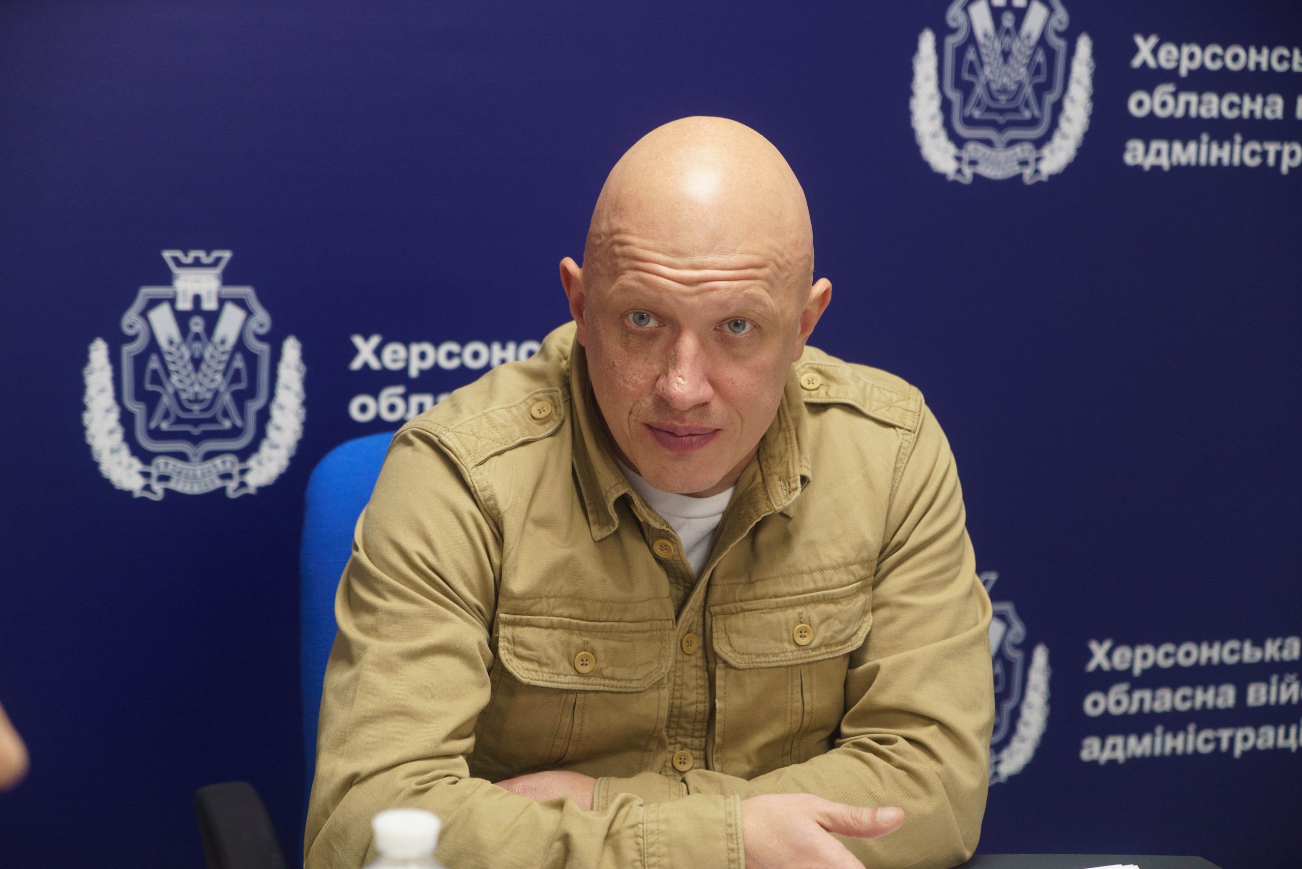 Чоловік у бежевій сорочці сидить за столом на фоні синього брендованого банера з логотипом Херсонської обласної військової адміністрації. Він склав руки перед собою та серйозно дивиться в об'єктив камери під час офіційного заходу.