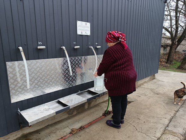 Жінка набирає воду у велику пластикову пляшку зі спеціально обладнаного пункту розливу води. Пункт має кілька кранів, виведених через металеву стіну технічної споруди.