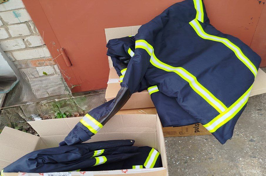 Комплекти спеціального захисного одягу темно-синього кольору зі світловідбивними жовтими смугами. Одяг лежить на картонних коробках поруч із цегляною будівлею.