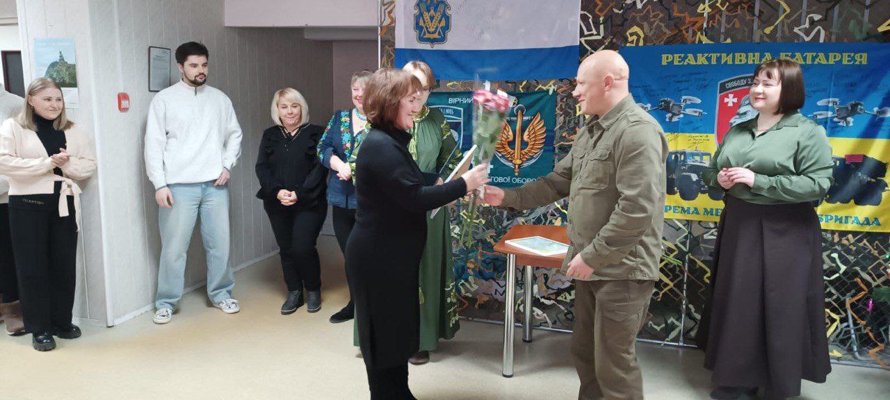 Урочиста мить у приміщенні з державною та військовою символікою: чоловік у камуфляжній формі вручає букет квітів і грамоту жінці. На фоні за ними спостерігають інші учасники події, а поруч розміщено банери з написами: Реактивна батарея та зображеннями військової техніки.