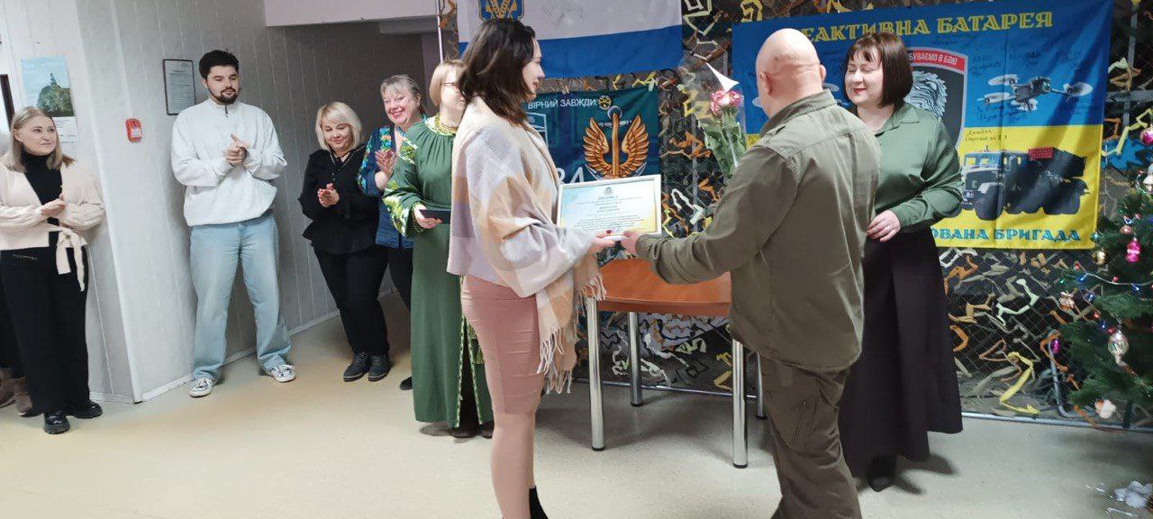 Урочиста подія у приміщенні з військовою символікою: чоловік у камуфляжній формі вручає почесну грамоту та квіти жінці. На фоні присутні горі гості, які аплодують, а праворуч стоїть прикрашена новорічна ялинка та розміщені тематичні банери Реактивна батарея.