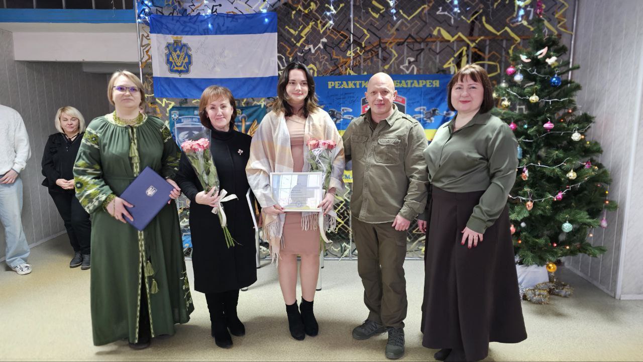 Група людей стоїть у приміщенні поруч із прикрашеною новорічною ялинкою під час урочистої події. У центрі жінка тримає почесну грамоту та квіти, а інші учасники тримають нагороди та букети на фоні прапорів із військовою символікою.