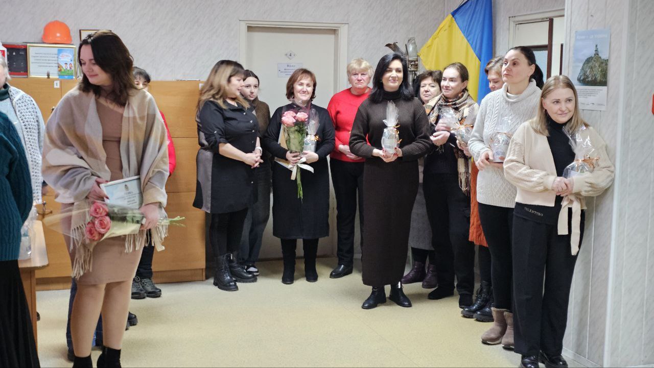 Група жінок стоїть у приміщенні під час урочистого заходу. Деякі з них тримають у руках квіти, грамоти та невеликі подарунки у прозорому пакуванні. На фоні за ними видно державний прапор України та частину інтер'єру кабінету.
