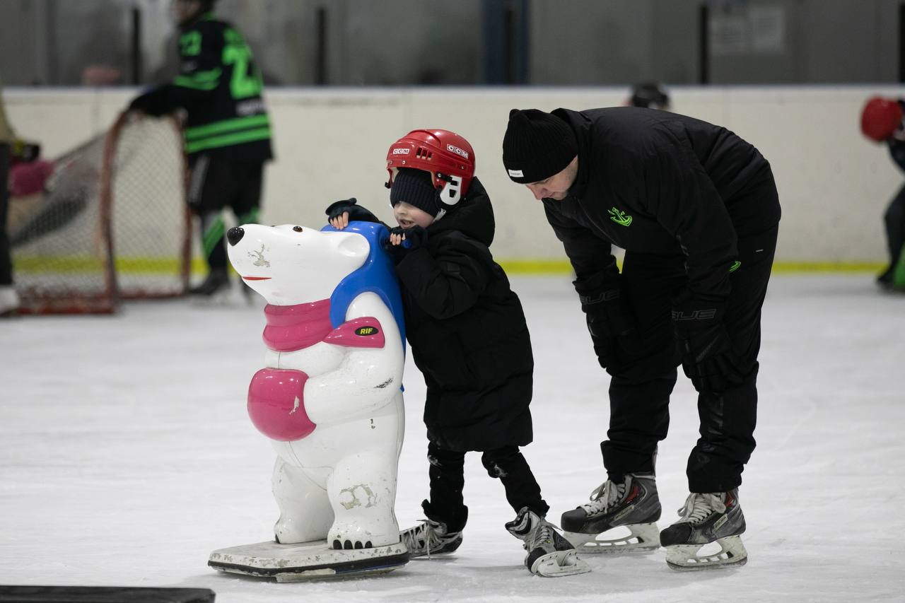 На льодовій арені чоловік у чорному спортивному костюмі допомагає дитині в червоному шоломі та чорній куртці вчитися кататися на ковзанах. Дитина тримається за спеціальний тренажер-підтримку у формі білого ведмедя з рожевим шарфом. На задньому плані розмито видно іншого гравця біля хокейних воріт.