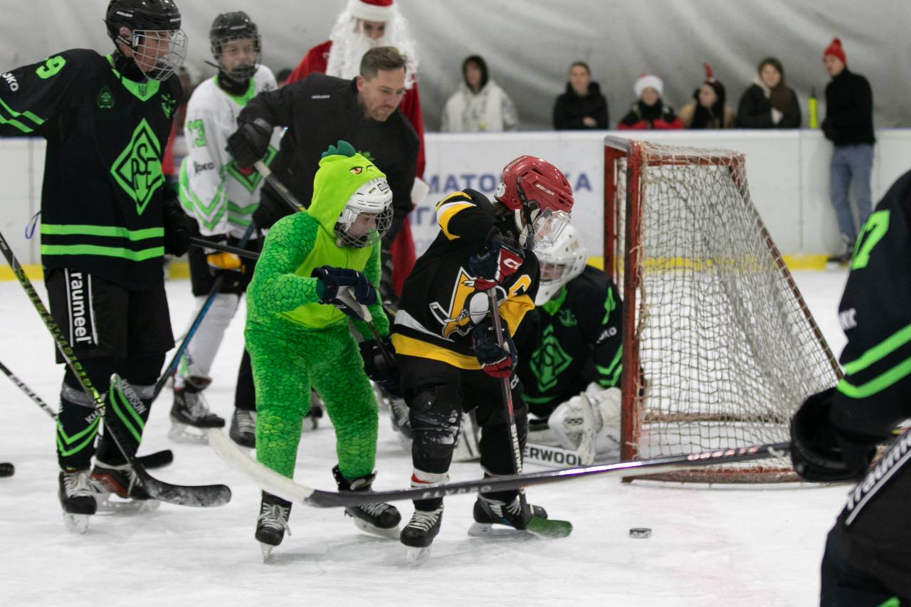 Динамічна сцена хокейного матчу на льодовій арені. У центрі кадру — юний гравець у яскравому зеленому костюмі динозавра та інший гравець у чорно-жовтій формі борються за шайбу біля воріт. На задньому плані видно дорослих гравців, суддів та глядачів, серед яких помітно аніматора у костюмі Святого Миколая.