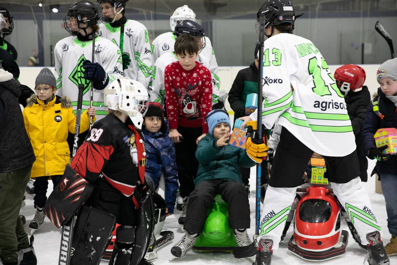 На льодовій арені хокеїст у біло-зеленій формі передає подарунок дитині, яка сидить на спеціальній пластиковій фігурі для катання. Навколо знаходяться інші гравці команди в екіпіруванні та діти у зимовому одязі, деякі з них тримають такі самі яскраві подарункові коробки.