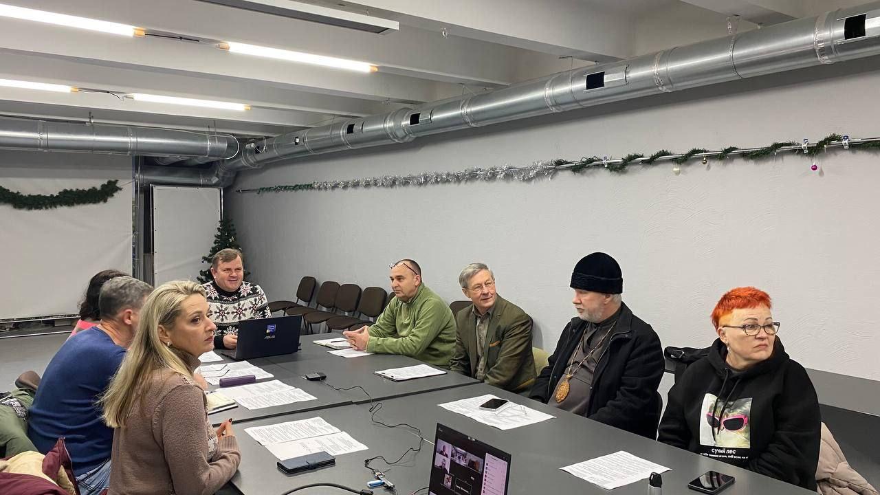 Група людей сидить за великим столом під час ділової зустрічі або наради в сучасному приміщенні з новорічним декором. На столі лежать документи та ноутбуки, учасники залучені в обговорення.