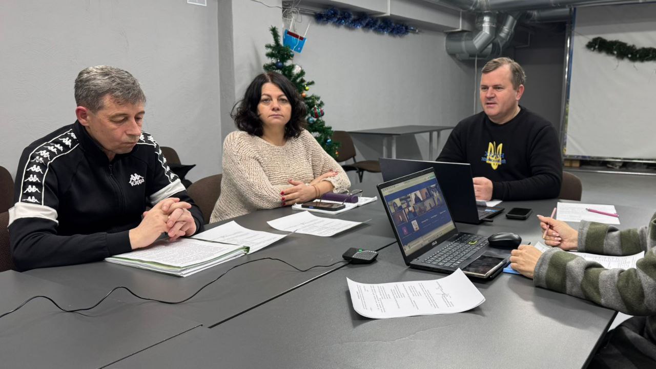 Група людей сидить за великим столом під час робочої зустрічі в приміщенні з новорічним декором. На столі лежать документи, ноутбуки та телефони; на екрані одного з ноутбуків видно вікно відеоконференції.