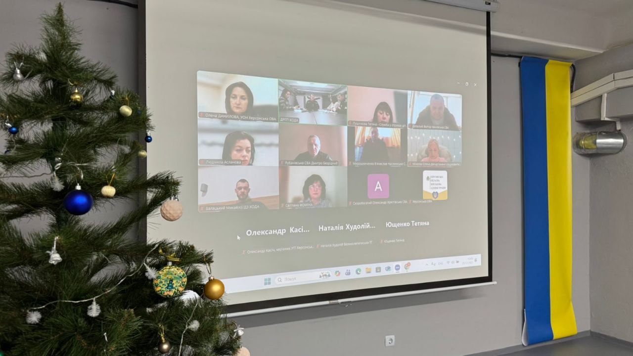 Проєкційний екран у приміщенні, на якому відображено онлайн-нараду у форматі відеоконференції з багатьма учасниками. Поруч із екраном стоїть прикрашена новорічна ялинка, а на стіні праворуч розміщено синьо-жовтий прапор.
