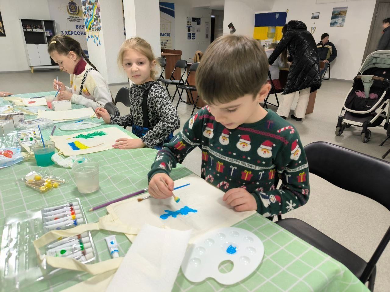 Діти займаються творчістю під час майстер-класу в просторому світлому залі. На передньому плані хлопчик у святковому светрі наносить блакитну фарбу на екосумку. Поруч дві дівчинки також розмальовують свої сумки; на одній із них уже зображена зелена ялинка над синьо-жовтим прапором. На столі, застеленому зеленою скатертиною, розкладені палітри, набори фарб та склянки з водою.