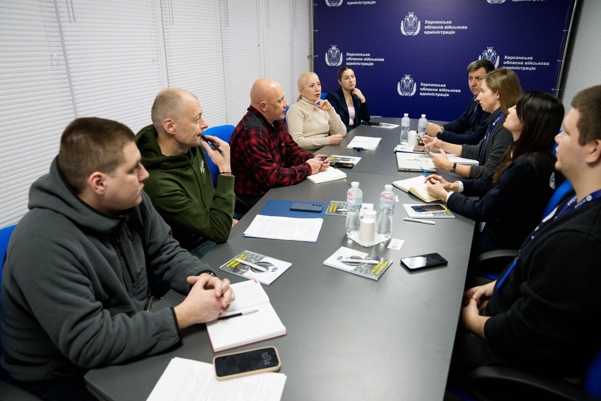 Робоча зустріч у приміщенні Херсонської обласної військової адміністрації. Група людей сидить за довгим столом під час обговорення. Учасники мають при собі блокноти для записів, телефони та воду. На фоні — синій банер із логотипом адміністрації, вікна зачинені жалюзі.