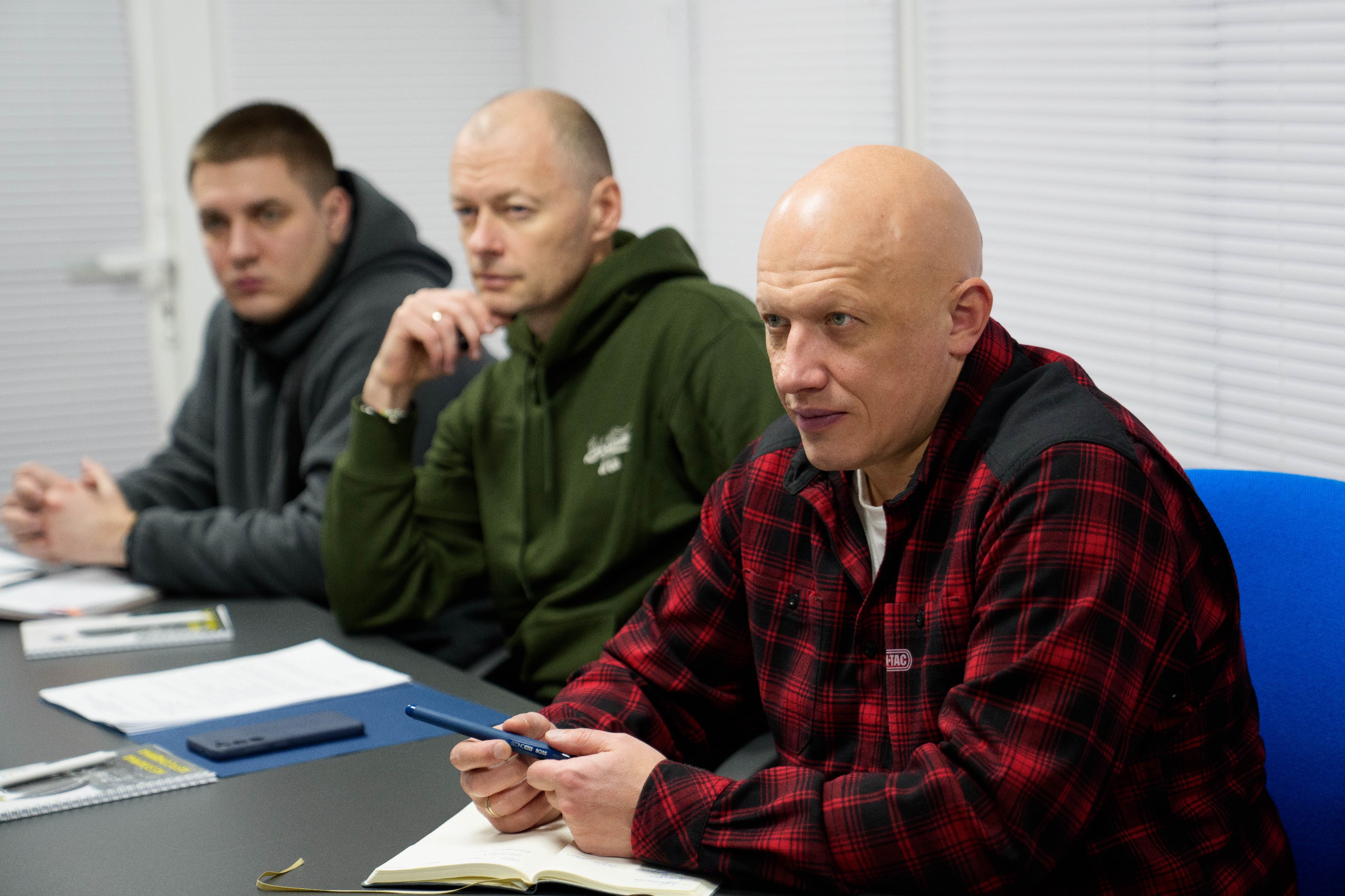 Учасники робочої наради в офісному приміщенні. Троє чоловіків сидять за столом, уважно слухаючи доповідача. Перед ними лежать відкриті блокноти для нотаток та ручки. На задньому плані видно білі жалюзі на вікнах.