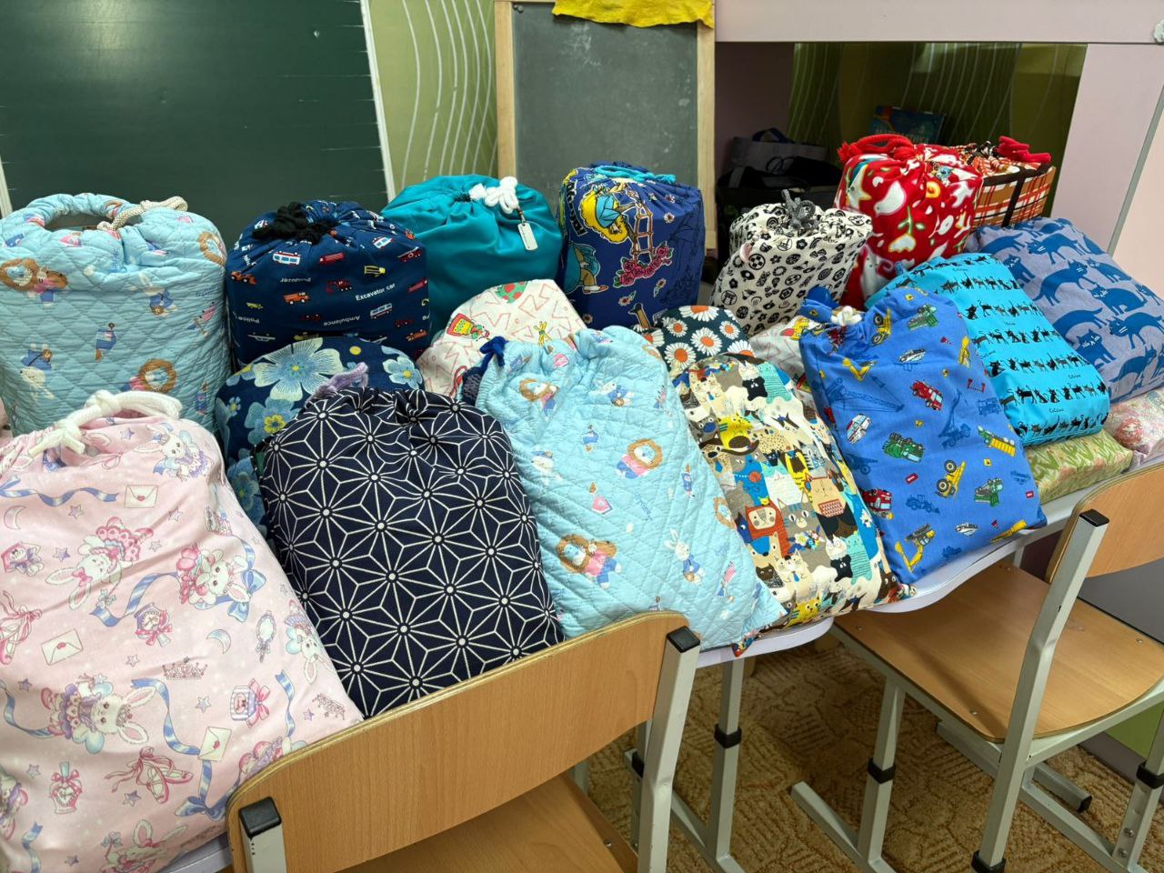 Яскраві текстильні мішечки з подарунками для дітей. Пакунки різних кольорів та з різноманітними принтами (квіти, машинки, тварини) щільно викладені на шкільних партах у світлій кімнаті. На задньому плані видно частину дошки та дитячі меблі.