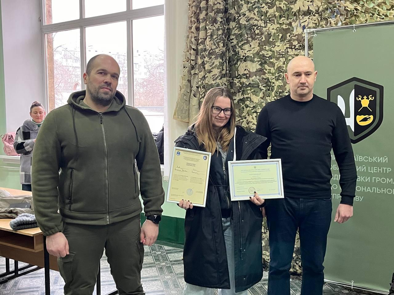 Урочисте вручення сертифікатів. Двоє чоловіків та жінка стоять у приміщенні на фоні стіни з маскувальною сіткою та інформаційним стендом. Жінка в центрі тримає в руках два рамкові документи. Учасники одягнені в повсякденний та тактичний одяг, атмосфера офіційна.