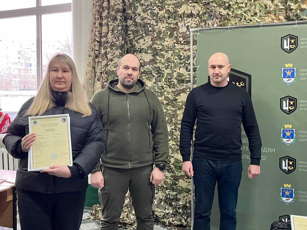 Вручення сертифікатів в офіційній установі. Чоловік у тактичному одязі та чоловік у чорному светрі стоять поруч із жінкою, яка тримає в руках рамковий документ. Позаду них розміщено зелений інформаційний стенд із символікою та стіну, декоровану маскувальною сіткою. На фоні через вікно видно зимовий міський пейзаж.