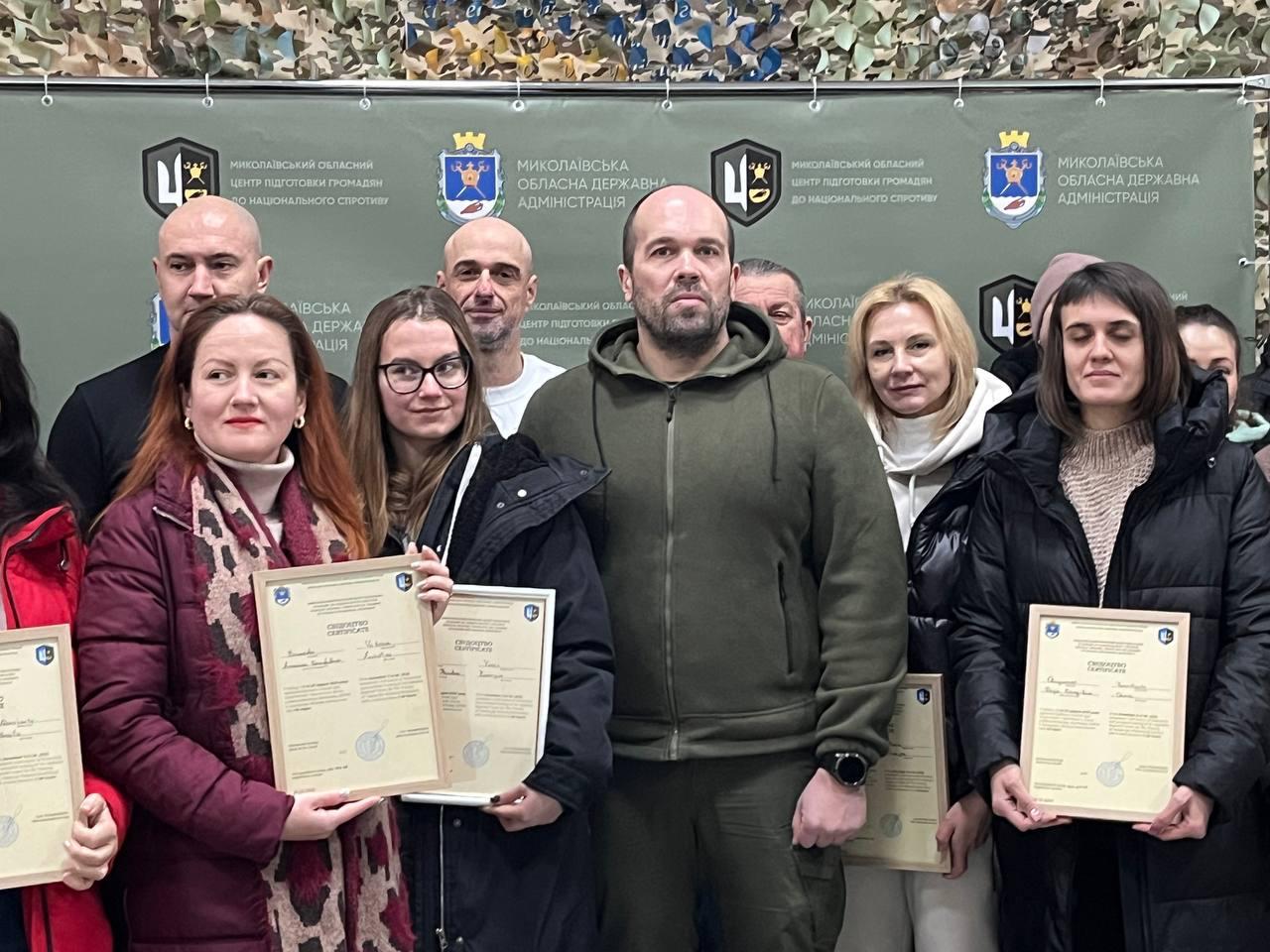 Урочисте вручення сертифікатів групі людей. У центрі кадру стоять чоловік у тактичному одязі та жінки, які тримають у руках рамкові документи. На задньому плані стіна, завішена маскувальною сіткою.