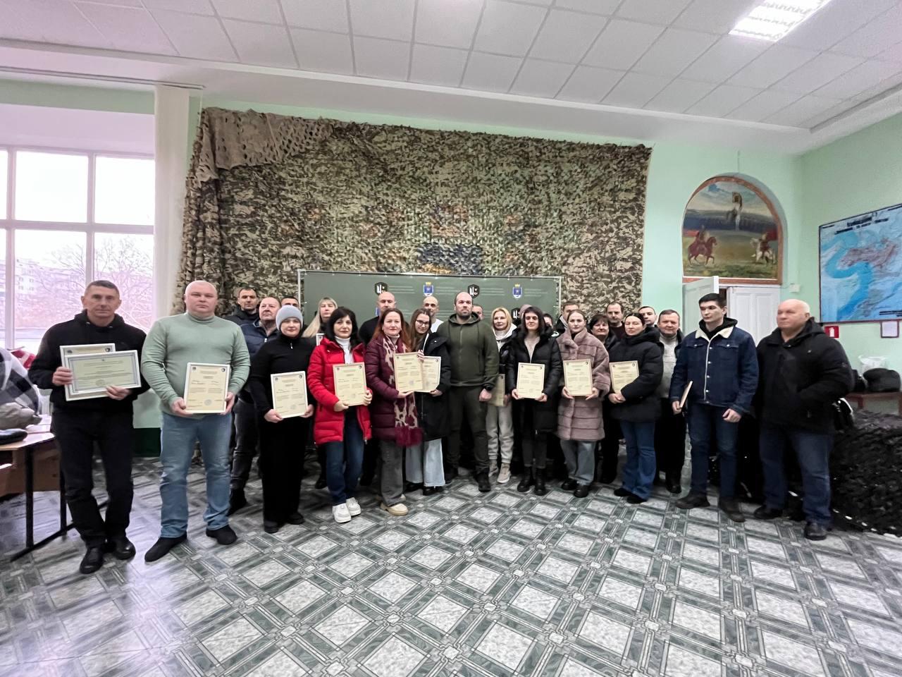 Групове фото учасників офіційного заходу в приміщенні. Велика група чоловіків та жінок стоїть у світлій залі, деякі з них тримають у руках урочисті грамоти або сертифікати. На задньому плані стіна частково завішена маскувальною сіткою, також видно інформаційні стенди та мапи.