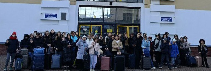 Група дітей та дорослих із валізами біля входу до залізничного вокзалу. Велика кількість людей у зимовому одязі зібралася на пероні перед скляними дверима касового залу. Дехто махає рукою на прощання або в знак вітання. Поруч із учасниками стоїть багато дорожніх сумок та чемоданів різних розмірів.