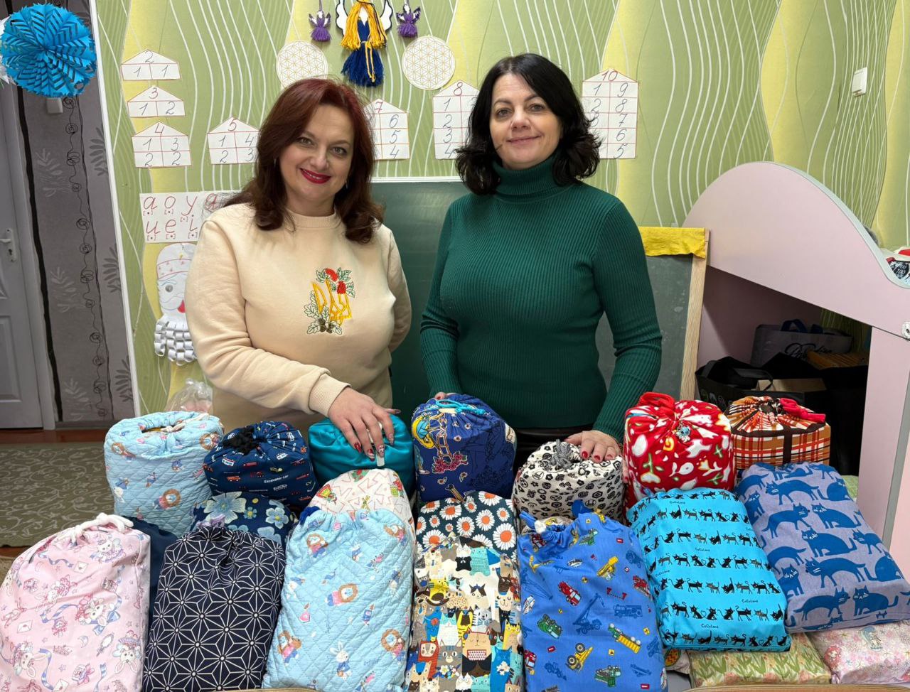 Дві жінки стоять біля столу з численними подарунками. Пакунки мають вигляд яскравих текстильних мішечків із різноманітними візерунками. Жінки посміхаються, поклавши руки на подарунки. На задньому плані видно зелену стіну класу з навчальними матеріалами та дитячими виробами.