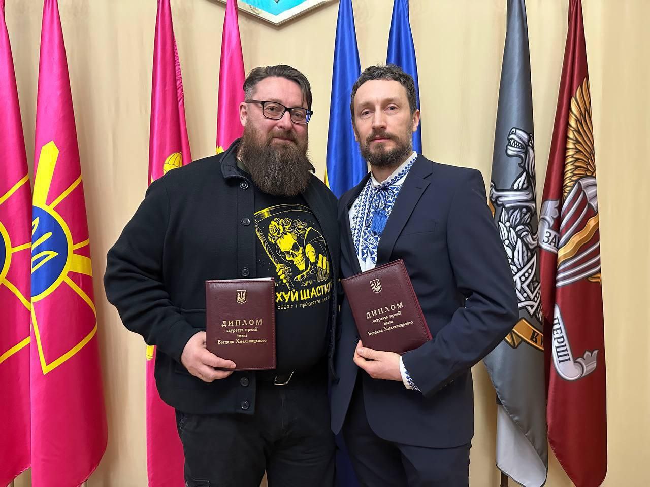Двоє чоловіків в офіційному вбранні тримають дипломи лауреатів премії імені Богдана Хмельницького. Вони стоять поруч на фоні державних та військових прапорів України. Один чоловік одягнений у чорну куртку та футболку з принтом, інший — у класичний костюм та вишиванку.