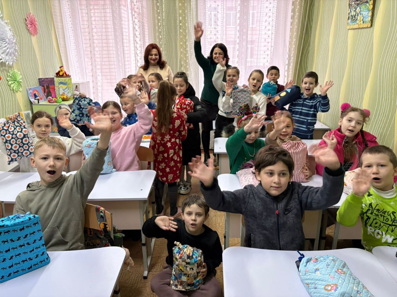 Група школярів у класі радісно махають руками на камеру. Діти сидять за партами, тримаючи кольорові святкові пакунки. На задньому плані стоять двоє вчительок, які також посміхаються. Кімната прикрашена дитячими малюнками та паперовими виробами.