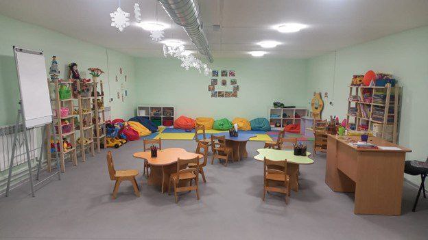 Інтер'єр просторої та світлої дитячої кімнати або ігрової зони. У приміщенні розставлені маленькі дерев'яні столи та стільці, стелажі з іграшками та книгами, а вздовж стіни розміщені різнокольорові м'які крісла-мішки.