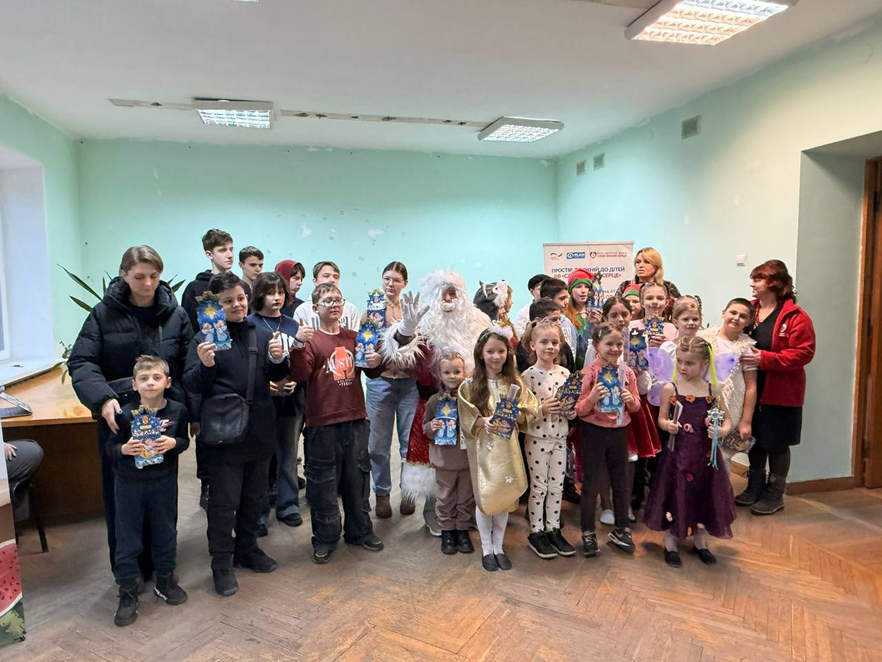 Група дітей та дорослих у приміщенні під час святкового заходу. У центрі стоїть аніматор у костюмі Санта-Клауса (або Святого Миколая). Деякі діти вдягнені у святкові костюми янголів та фей, інші тримають у руках подарунки у синіх пакунках.