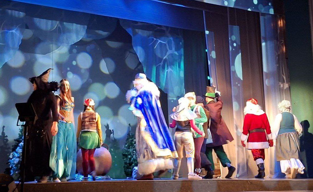 На сцені театру проходить святкова вистава за участю багатьох персонажів у яскравих костюмах. Серед героїв — Святий Миколай у синій мантії, Санта-Клаус у традиційному червоному вбранні, а також персонажі в казкових строях. Сцену прикрашено атмосферною підсвіткою з ефектом мерехтіння на фоні зимових декорацій.