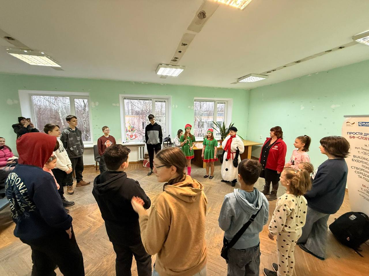 Група дітей та дорослих стоїть колом у світлій кімнаті під час святкового заходу. У центрі кола стоять троє дітей у карнавальних костюмах — двоє ельфів та сніговик. Присутні спостерігають за виступом або готуються до спільної гри.