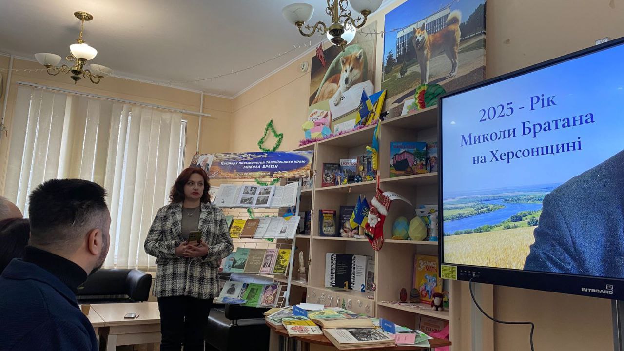 Фото з бібліотечного заходу, присвяченого Року Миколи Братана на Херсонщині. У залі перед слухачами виступає жінка, поруч стоїть книжкова виставка з творами письменника та великий екран із тематичним написом і пейзажем Херсонщини.