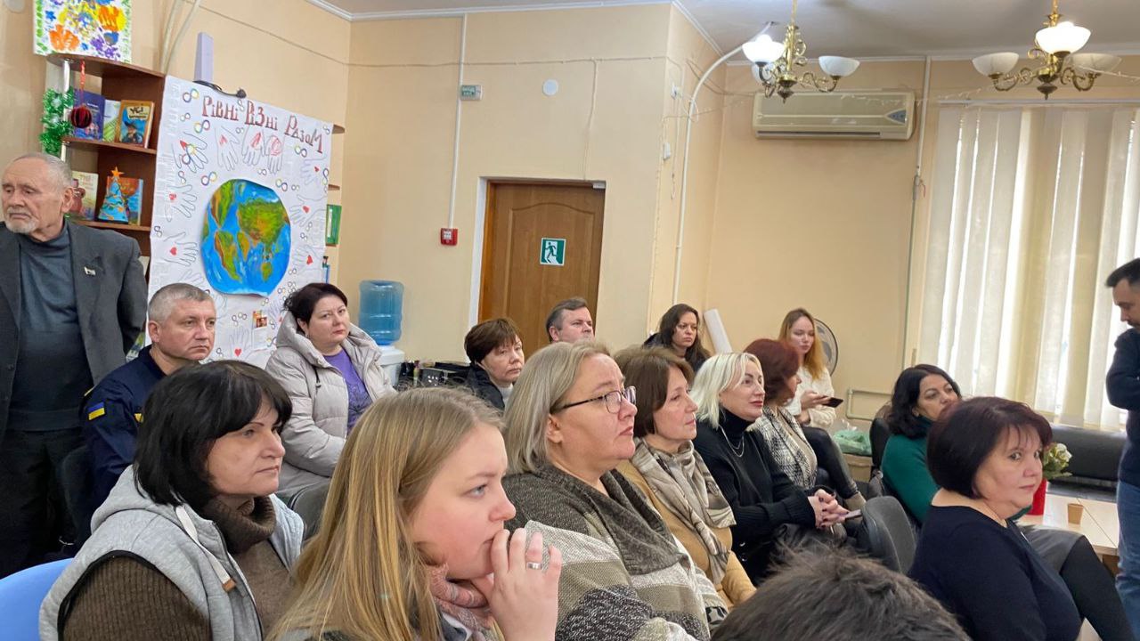 Фото з культурного заходу в бібліотеці. У залі сидять слухачі, які уважно спостерігають за виступом. На стіні розміщено плакат із зображенням земної кулі та гаслом «Рівні. Різні. Разом», а в кутку зали видно книжкові полиці та елементи святкового декору.