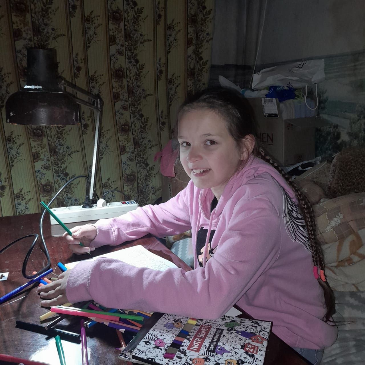 Дівчинка сидить за столом і займається творчістю — малює олівцями та фломастерами. Вона одягнена у рожеве худі, а її волосся заплетене в довгу косу. На столі лежать розмальовка, блокнот та багато кольорових інструментів для малювання під світлом настільної лампи.