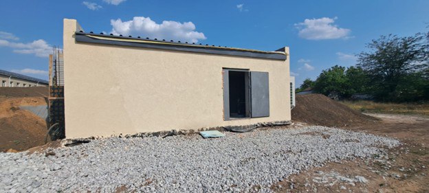 Зовнішній вигляд новозбудованої одноповерхової споруди (ймовірно, наземного укриття) з бежевим фасадом та масивними металевими дверима. Навколо будівлі тривають роботи з благоустрою, територія засипана щебенем.
