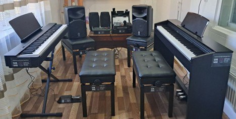 Комплект професійного музичного та звукового обладнання в інтер'єрі: два електронні піаніно з банкетками, акустичні системи, мікшерний пульт та мікрофон. Обладнання розміщене у світлій кімнаті з ламінатом на підлозі.