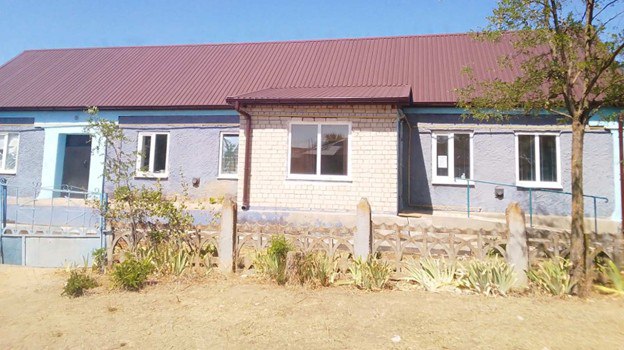 Одноповерхова будівля з блакитним фасадом, білою цегляною прибудовою та новим коричневим дахом із металочерепиці під ясним небом.