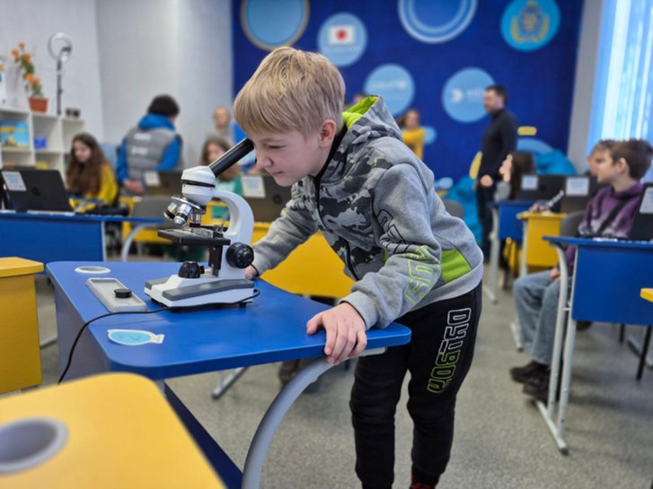 Учень молодших класів зосереджено дивиться у професійний мікроскоп під час практичного заняття. На фоні видно сучасний освітній простір з іншими дітьми та дорослими, оформлений у синьо-жовтих кольорах із логотипами партнерів.