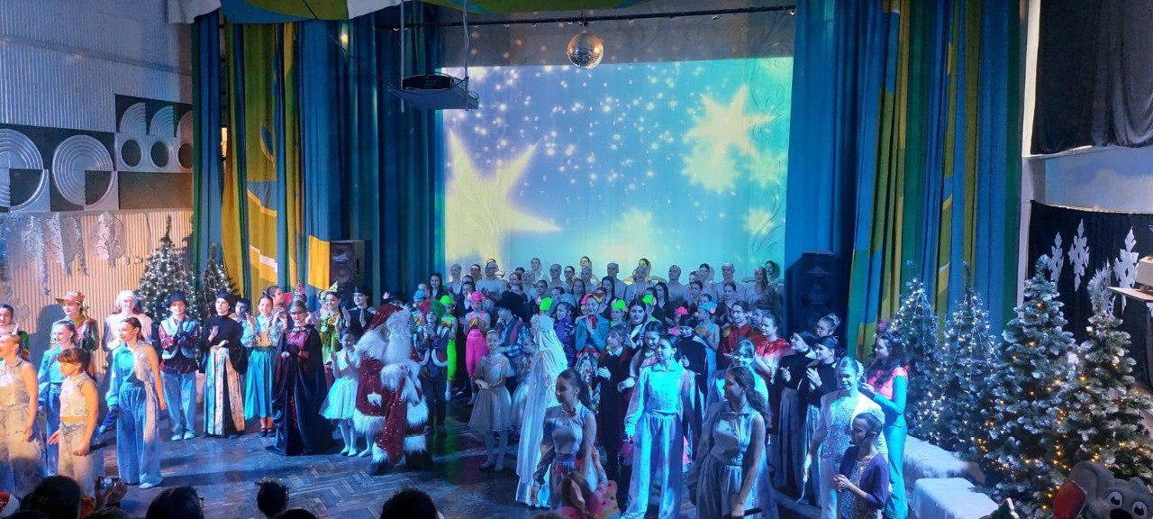 Велика група артистів у різноманітних сценічних костюмах, серед яких Святий Миколай, позує на сцені після завершення вистави. Задній план прикрашений великим екраном із зображенням зоряного неба, а з боків сцени стоять новорічні ялинки з гірляндами.