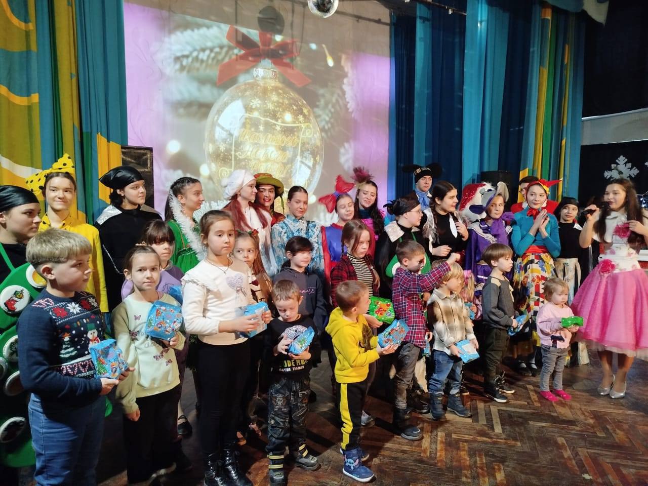 Велика група дітей та підлітків у яскравих костюмах стоїть на сцені. Дехто з дітей тримає в руках невеликі подарунки у синьому пакуванні. На задньому плані — екран із зображенням новорічної кульки, а підлога сцени дерев'яна.
