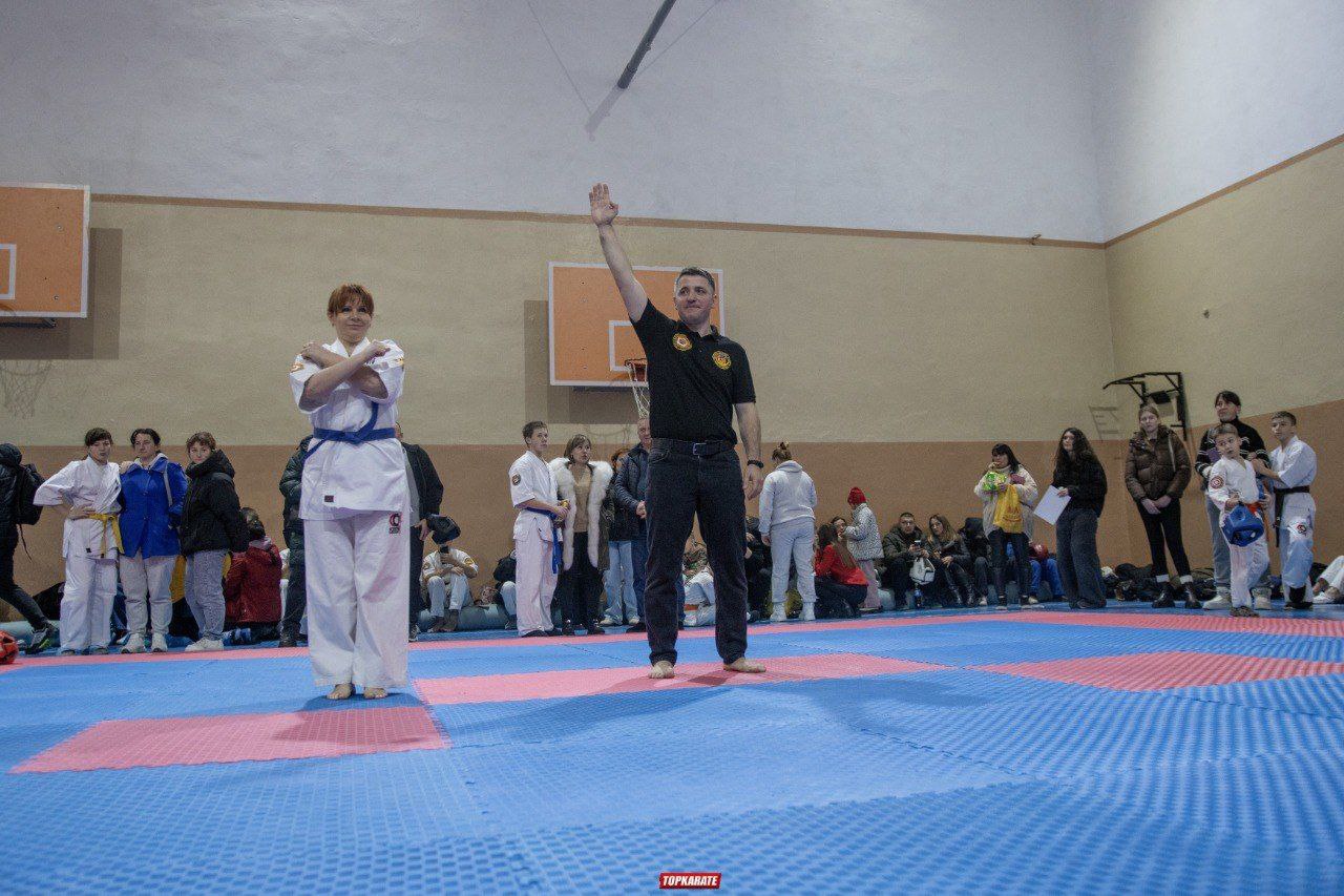 Спортивний суддя, який підняв праву руку догори та спортсменка, яка склала руки хрест-навхрест, стоять на синьо-рожевому татамі в спортивній залі; на задньому плані - глядачі та інші учасники.