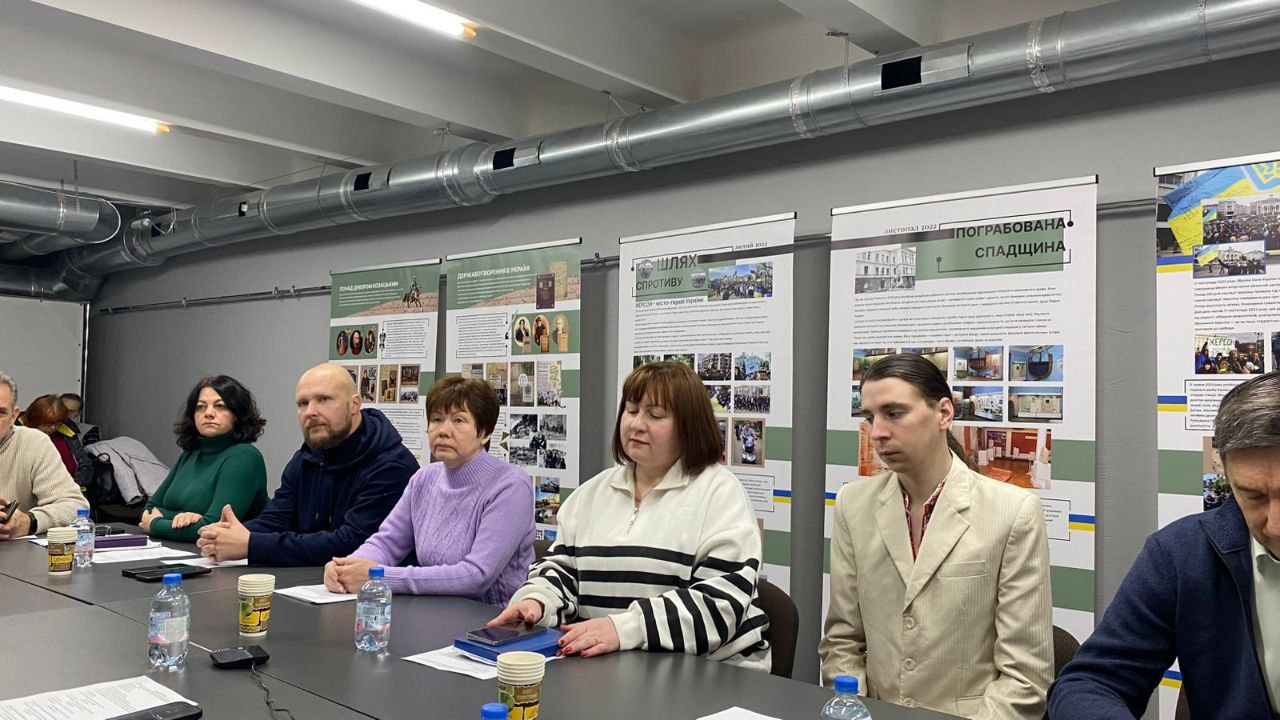 Учасники зустрічі сидять за столом у сучасному залі; на задньому плані розміщені інформаційні банери про історію та спротив Херсонщини, зокрема з назвами: Шлях спротиву та Пограбована спадщина.