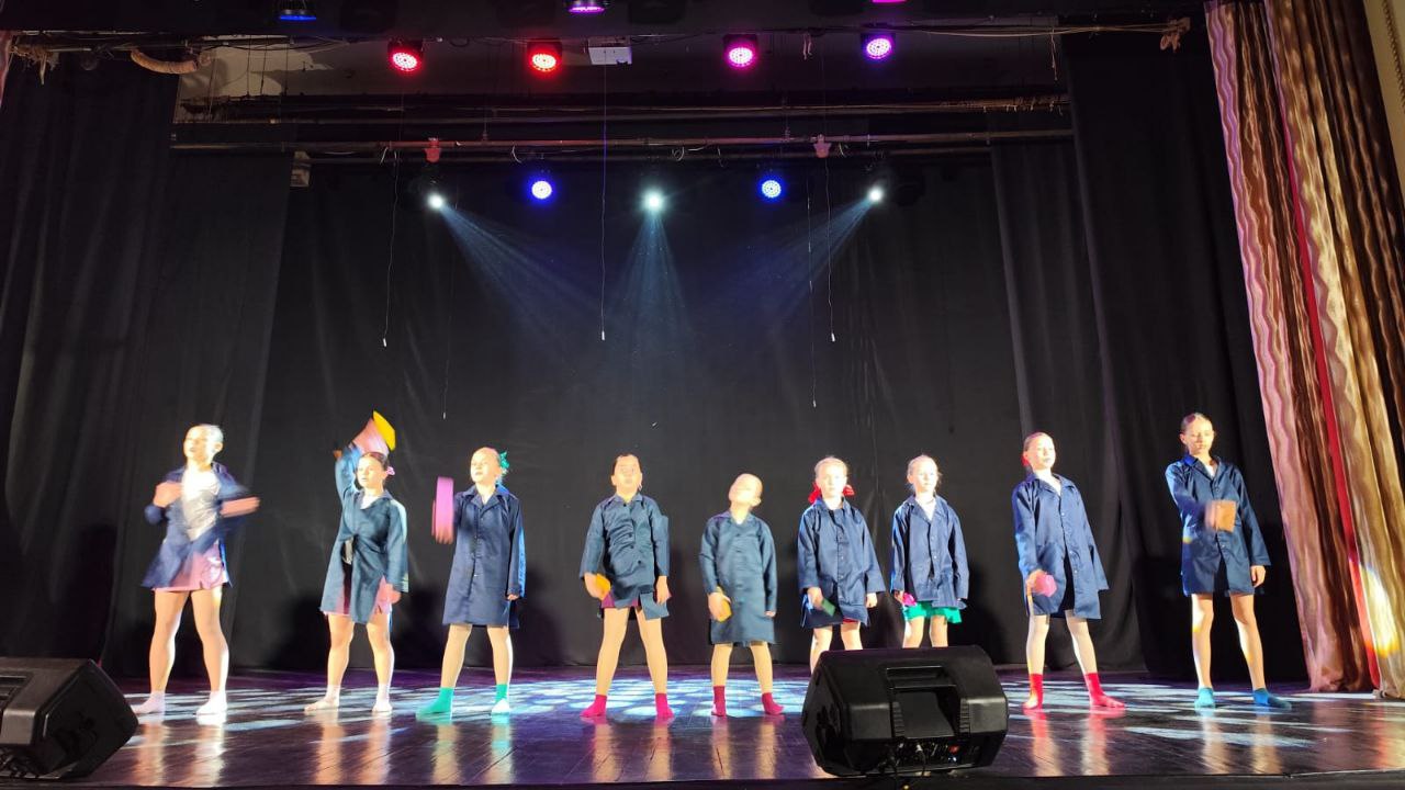 Дев'ять дівчаток стоять на сцені перед чорною завісою, освітлені різнокольоровими прожекторами. Вони одягнені в довгі темно-сині сорочки або плащі та шорти, і деякі з них мають яскраві неонові шкарпетки або чобітки.