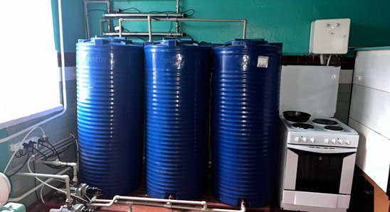 Три великі сині вертикальні баки для зберігання води стоять у куті кімнати поруч із білою електричною плитою, на тлі стін кольору морської хвилі.