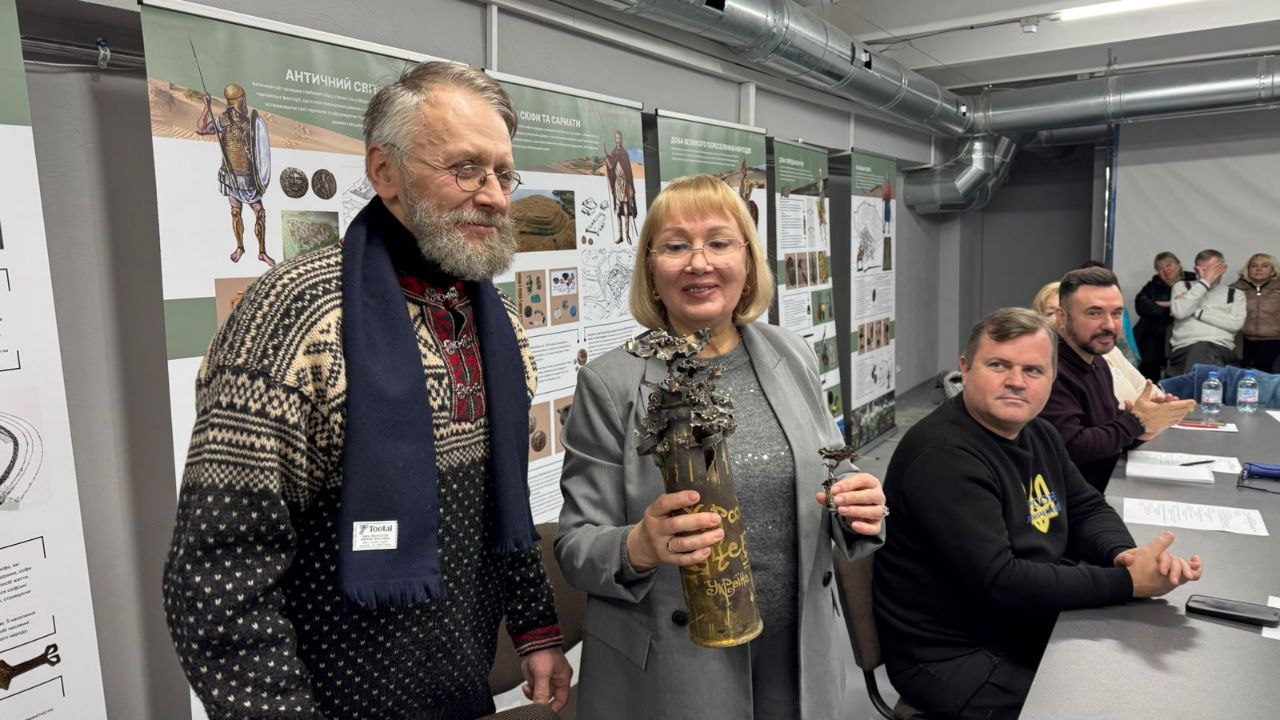 Чоловік у в'язаному светрі та жінка в сірому піджаку тримають у руках гільзу, декоровану металевими квітами, на фоні банерів історичної виставки.