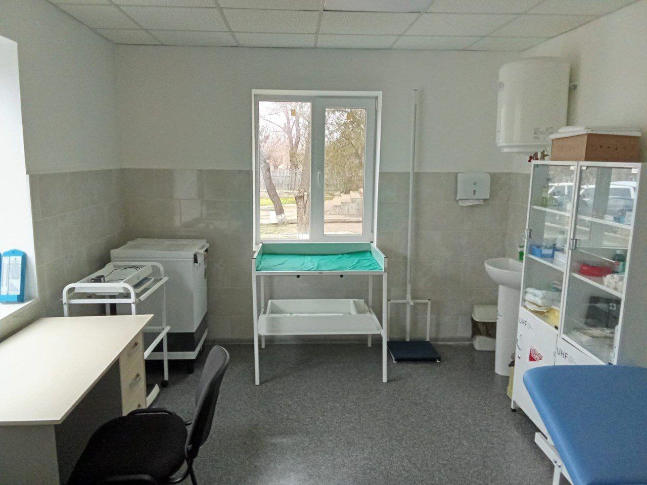 Інтер'єр сучасного оглядового кабінету: стіни облицьовані світлою плиткою, встановлено скляну шафу з медикаментами, ваги та стіл для огляду немовлят.