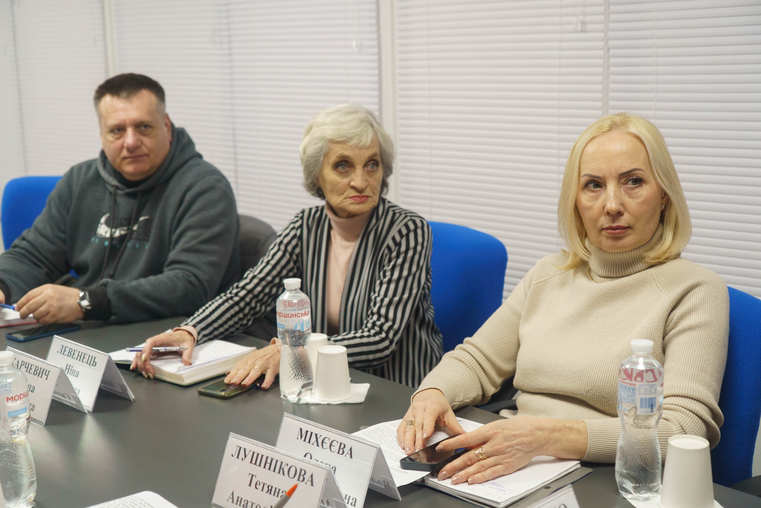 Троє учасників офіційної зустрічі сидять за робочим столом, уважно слухаючи доповідача. Перед ними розставлені таблички з іменами, блокноти для записів та пляшки з водою, а на фоні видно закриті білі жалюзі.