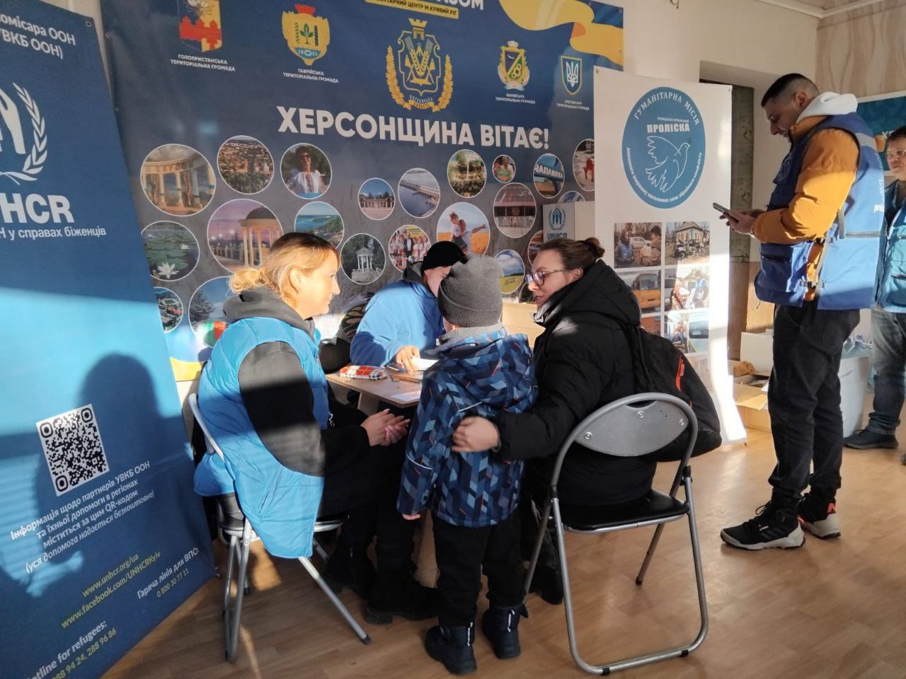 У приміщенні гуманітарного центру представники благодійних організацій спілкуються з жінкою та дитиною. На фоні розташований великий банер із написом «Херсонщина вітає!», гербами громад та логотипами міжнародних партнерів (УВКБ ООН, «Проліска»).