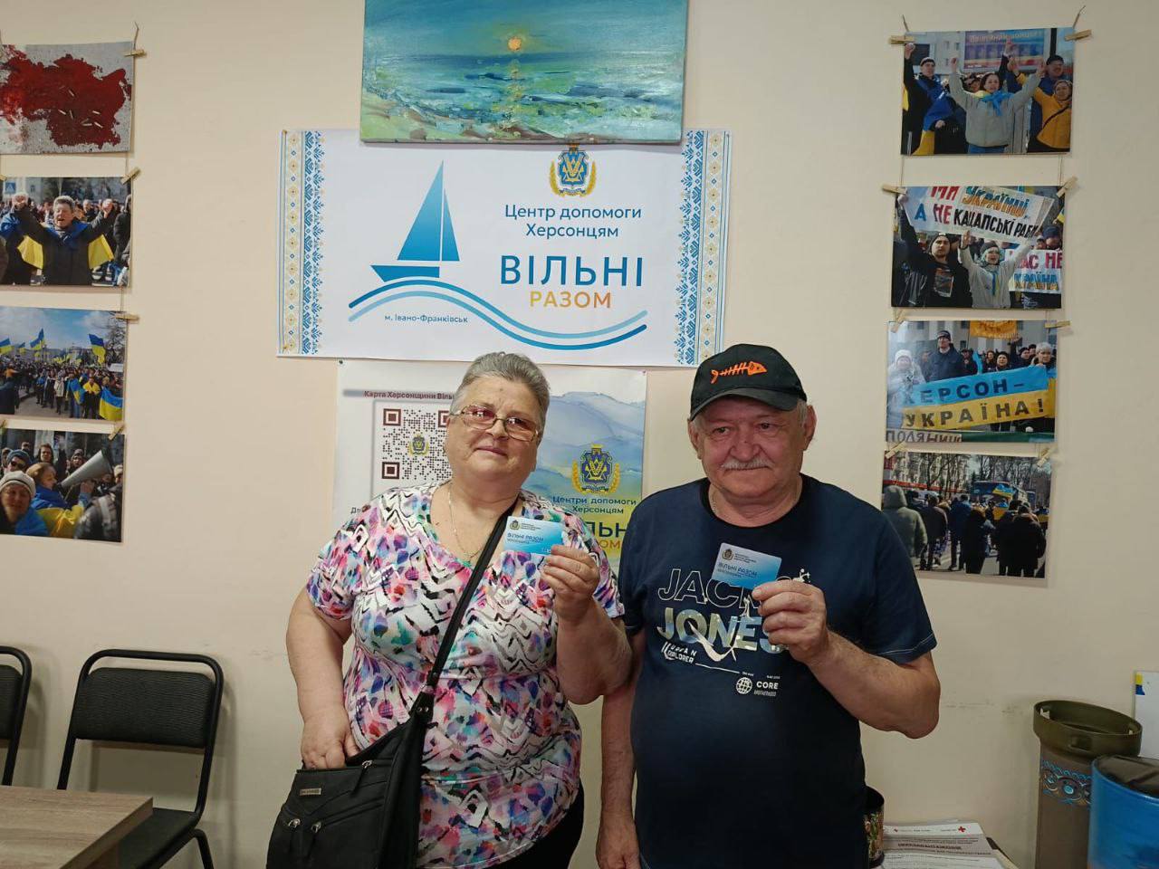 Чоловік та жінка стоять у приміщенні хабу та тримають у руках отримані посвідчення. На стіні за ними розміщено плакат «Центр допомоги херсонцям "Вільні разом"» та світлини з патріотичних мітингів.